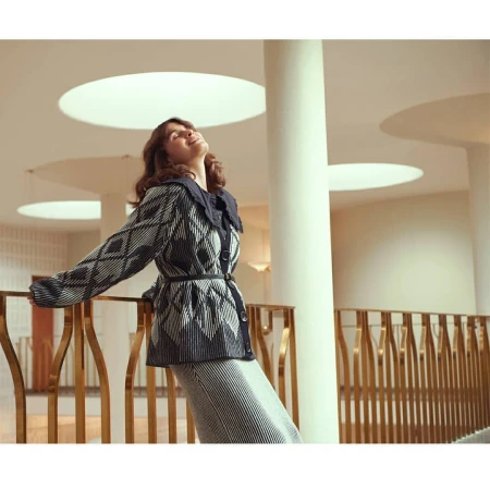 Woman in patterned cardigan and striped skirt poses indoors with head tilted back and eyes closed, near decorative railing and white pillar.