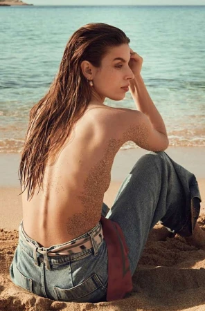 Topless woman with wet hair and sand on her back sitting on a sandy beach wearing denim trousers