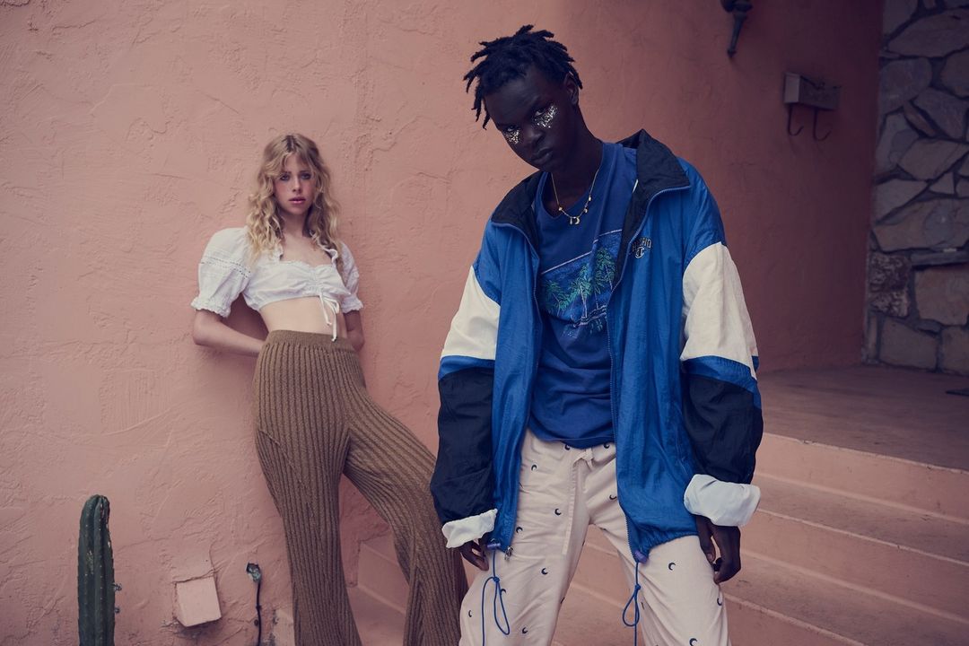 Two fashion models strike poses against a salmon-colored wall. A blonde woman wears a crop top and ribbed pants; a man wears a blue jacket.