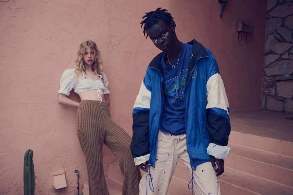Two fashion models strike poses against a salmon-colored wall. A blonde woman wears a crop top and ribbed pants; a man wears a blue jacket.