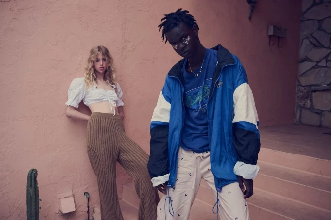 Two fashion models strike poses against a salmon-colored wall. A blonde woman wears a crop top and ribbed pants; a man wears a blue jacket.