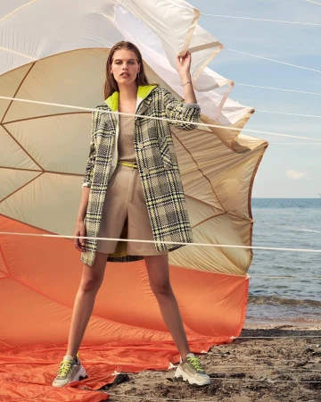 Model standing outdoors near a beach holding taut white fabric ropes in a plaid coat over beige shorts and sneakers.