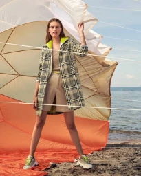 Model standing outdoors near a beach holding taut white fabric ropes in a plaid coat over beige shorts and sneakers.