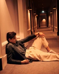 Man in dark blazer and light trousers seated on carpeted hotel corridor floor, leaning against wall