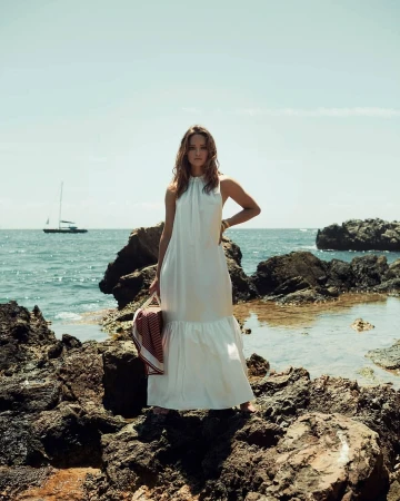Woman in a white sleeveless maxi dress standing on rocky coast holding a striped red towel with a sailboat on the ocean in the background
