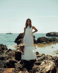 Woman in a white sleeveless maxi dress standing on rocky coast holding a striped red towel with a sailboat on the ocean in the background