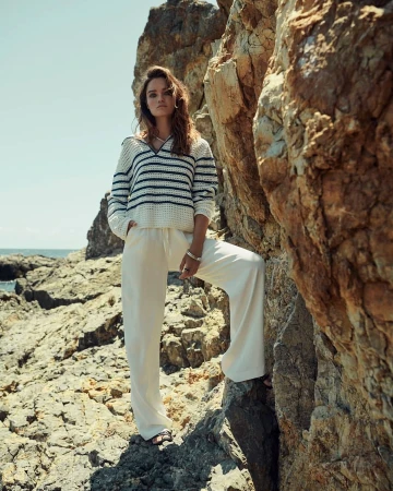 Woman wearing a striped sweater and white trousers posing on rocky terrain by the ocean by photographer Sune Czajkowski.