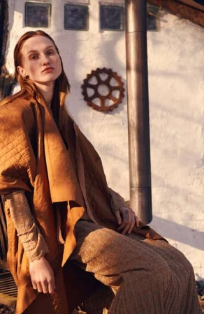 Model with red hair wearing a tan wrap coat and textured trousers seated outdoors against a textured white wall.
