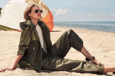 Woman in dark sunglasses wearing an olive green jacket and matching trousers sitting on a sandy beach.