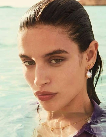 Close-up portrait of a woman with wet hair wearing pearl drop earrings and a delicate gold chain necklace in water.