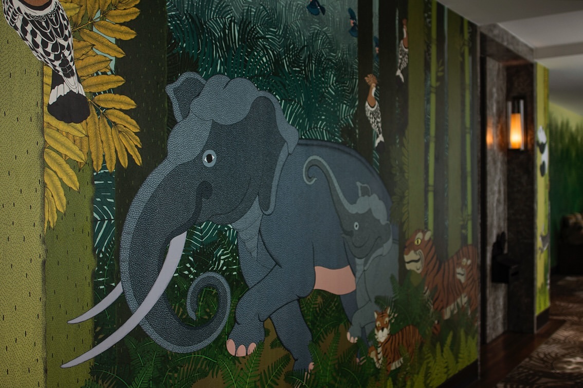 Illustration of a blue-gray elephant and baby elephant surrounded by tigers, birds, and foliage, on a wall with dark green background.