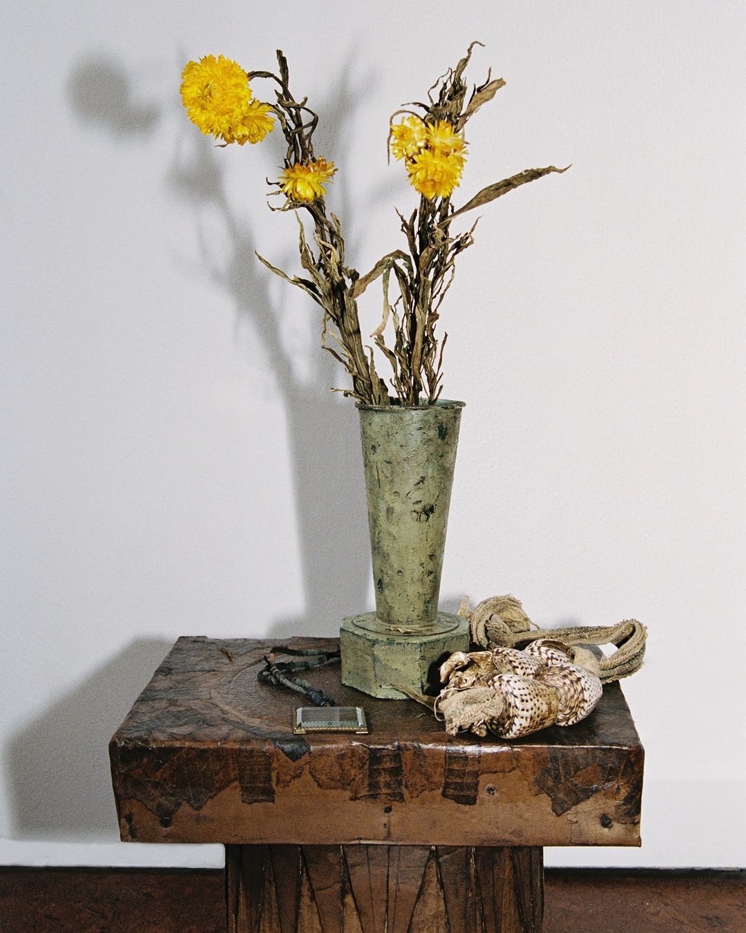 Still life photo of yellow flowers in green vase on carved wooden table with shells and beaded necklace in front of a white wall.