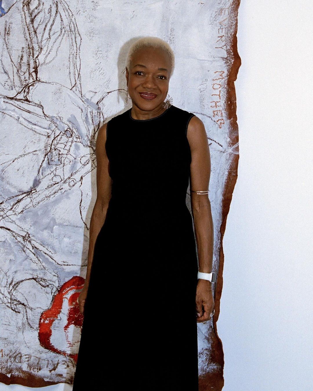 Woman in a black dress stands in front of an abstract art piece. The woman has short white hair, and neutral expression.