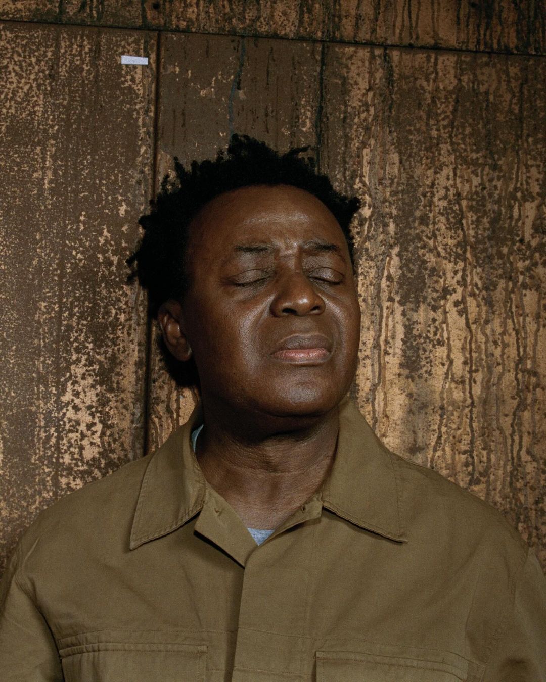 Portrait of an adult Black man with eyes closed, short black hair, wearing a brown collared shirt, against a brown textured background.