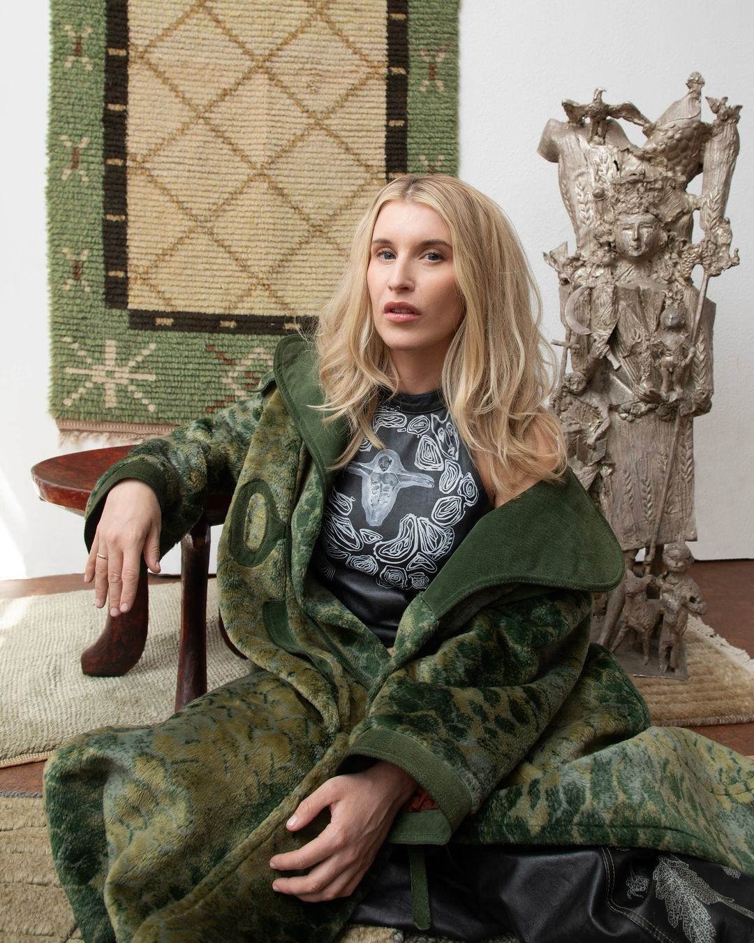 Woman in a patterned coat sits near a sculpted figure and woven wall hanging, creating a fashion portrait in an indoor setting.