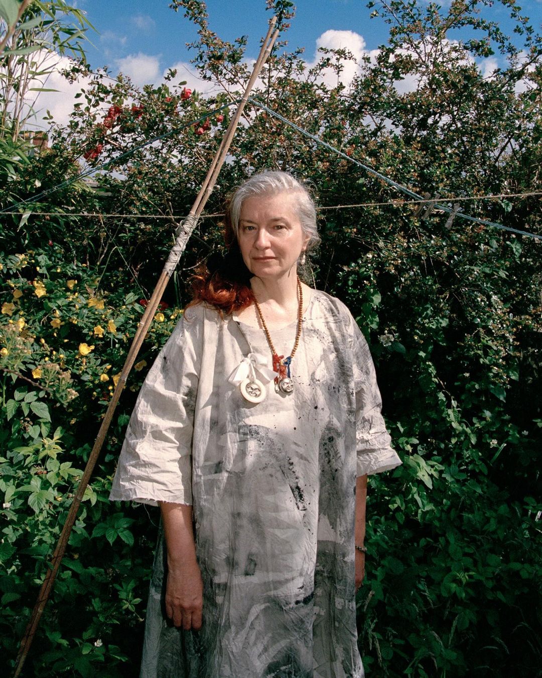 Portrait of an adult woman with long grey hair wearing a patterned dress, posing outdoors with trees and foliage in the background.
