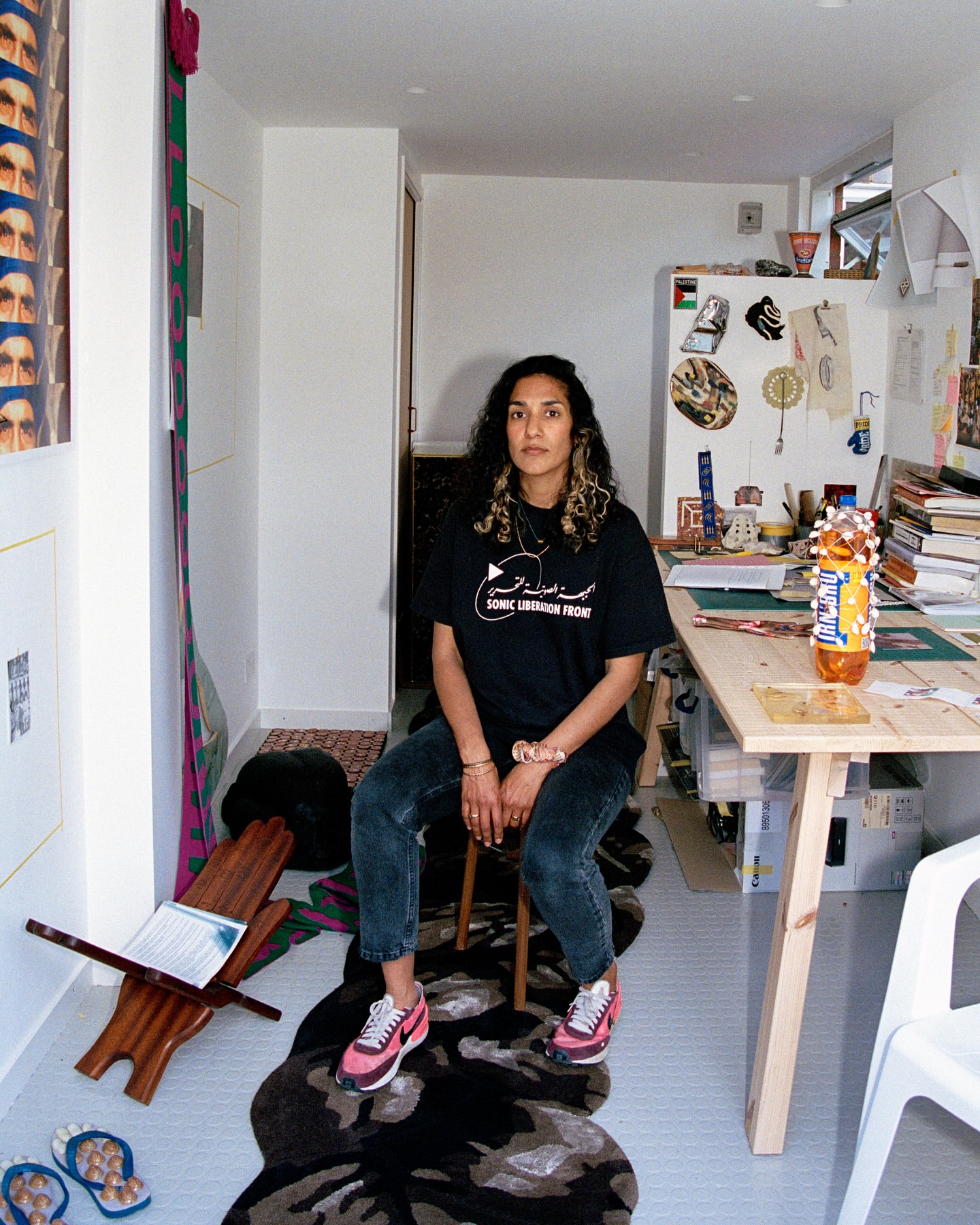 Portrait of a woman with curly hair sitting in her apartment studio, wearing a t-shirt with Arabic text, sneakers, jeans, and jewelry.
