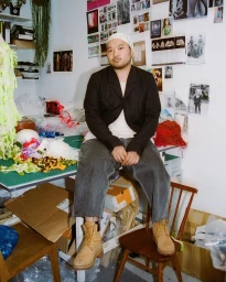 Man seated on a work table in a cluttered design studio wearing a black blazer and tan work boots