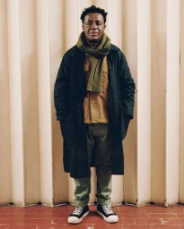 Portrait of a man wearing a dark coat, olive scarf, khaki jacket, and green trousers standing in London by Suzannah Pettigrew