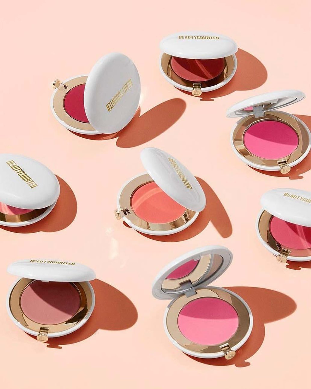 Several round, white and gold cosmetic compacts with pink and peach blush shades arranged on a soft peach background.