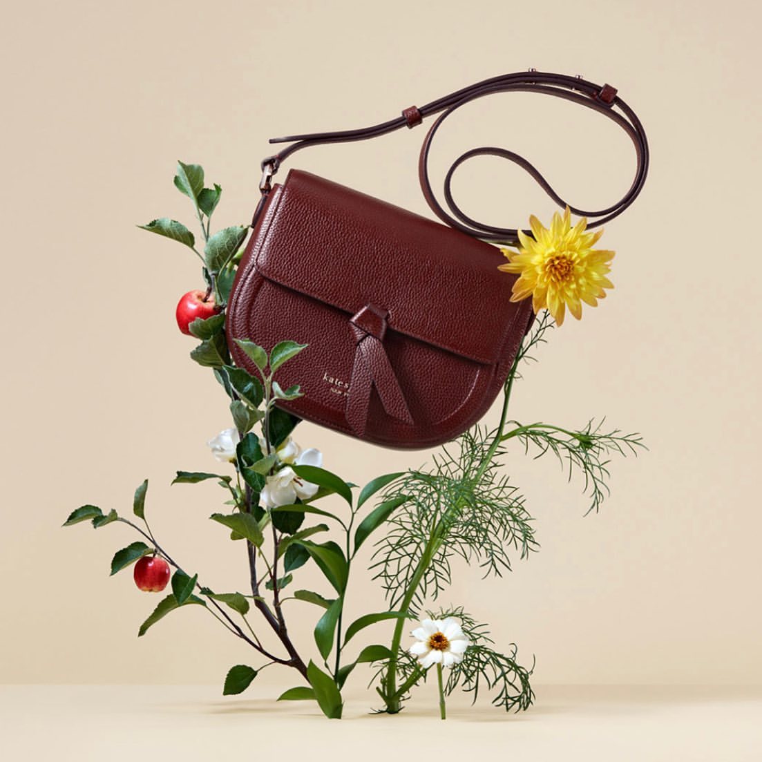 Burgundy leather purse with shoulder strap, displayed with apples, leaves, yellow and white flowers on a beige background.