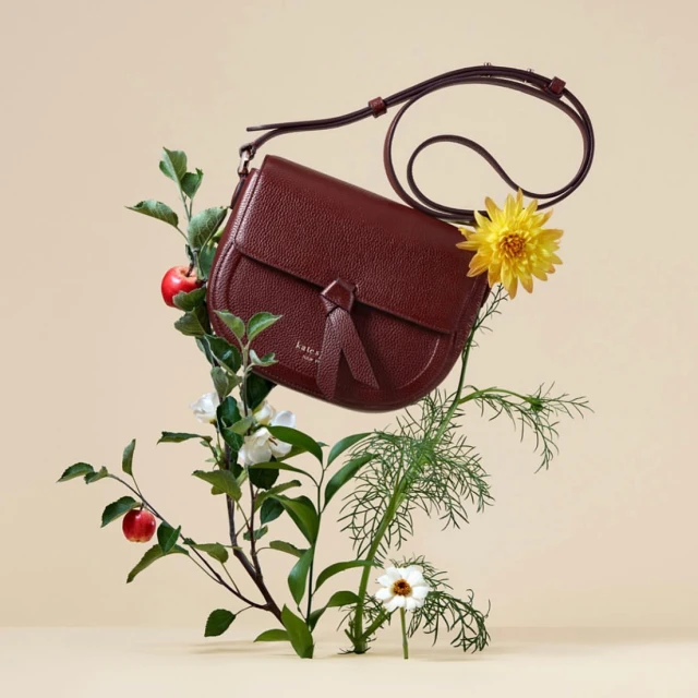 Burgundy leather purse with shoulder strap, displayed with apples, leaves, yellow and white flowers on a beige background.