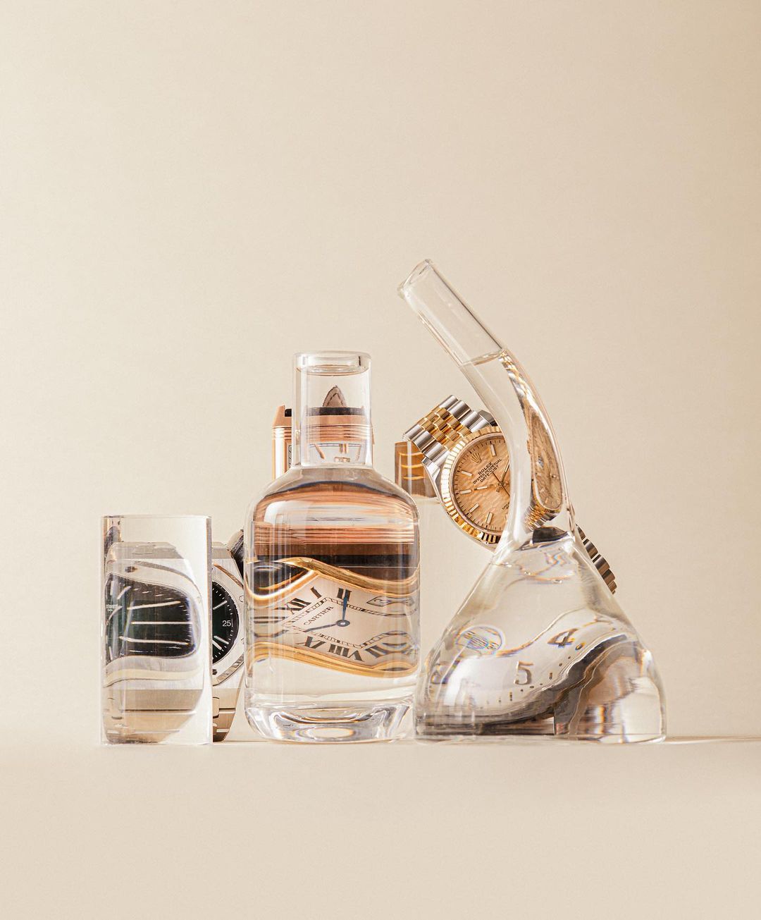 Still life studio photograph featuring wristwatches in clear liquid-filled glassware and a bottle against a neutral backdrop.