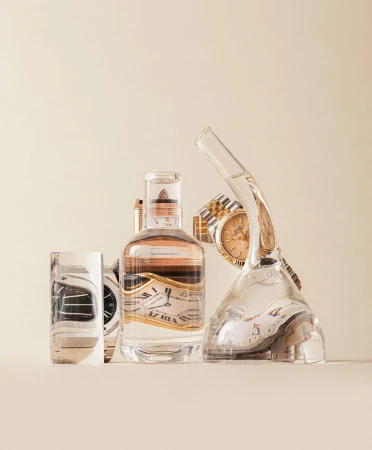 Still life studio photograph featuring wristwatches in clear liquid-filled glassware and a bottle against a neutral backdrop.