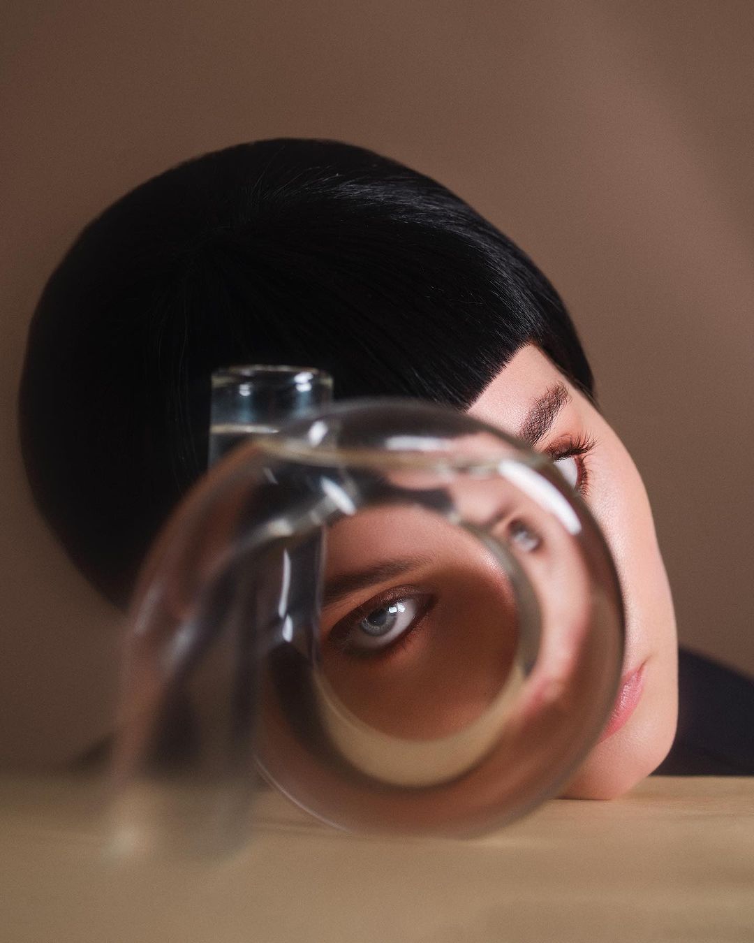 Portrait of a woman with dark hair and blue eyes looking through a glass with water, with a neutral background and a soft, diffused light.