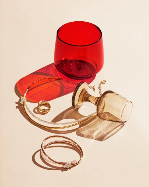 Still life studio shot of jewelry including a bracelet, ring, and necklace, arranged with two glasses casting bright shadows.