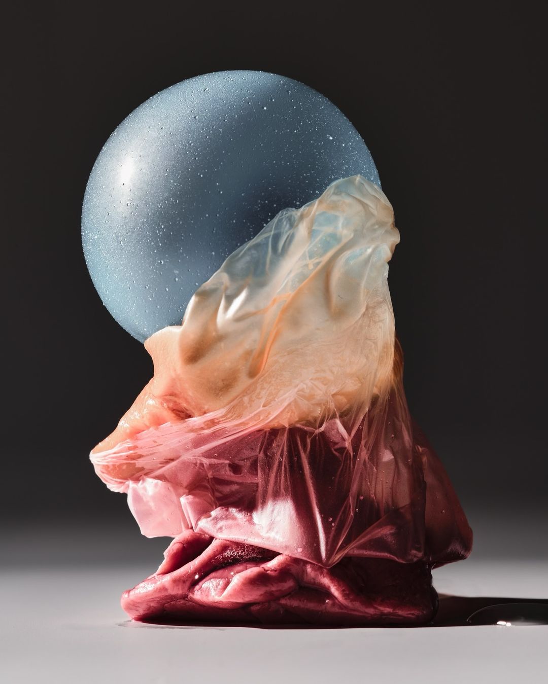 Surreal studio shot with a blue textured sphere atop layers of draped plastic sheeting in hues of pink, orange, and white against a dark background.
