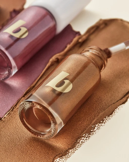 Close-up studio shot of bronze and maroon liquid shimmer product in clear dropper bottles with a gold logo against bronze makeup swatches.