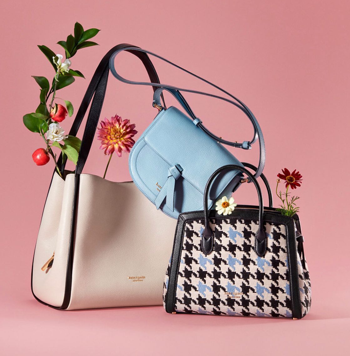 Three Kate Spade purses are showcased with floral accents against a pink backdrop in a studio product shot, highlighting fashion accessories.