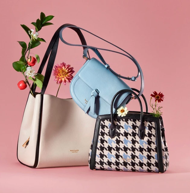 Three Kate Spade purses are showcased with floral accents against a pink backdrop in a studio product shot, highlighting fashion accessories.
