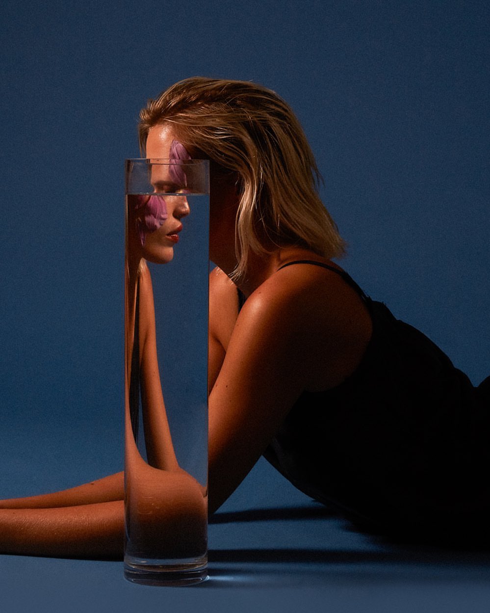 Portrait of a blond woman lying down with a clear glass vase on a blue background in a studio, capturing an artistic and stylish composition.