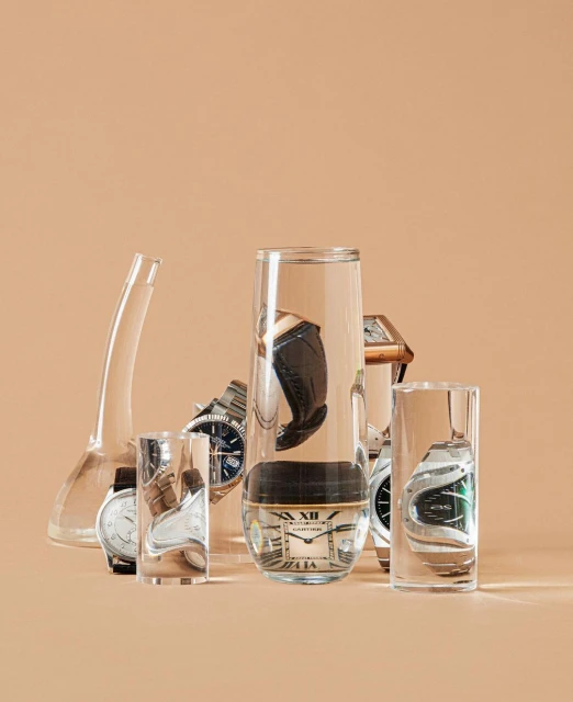 Conceptual studio shot of six wristwatches in glass containers against a neutral background. Watches have silver, gold, and leather bands.