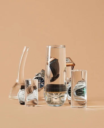 Conceptual studio shot of six wristwatches in glass containers against a neutral background. Watches have silver, gold, and leather bands.
