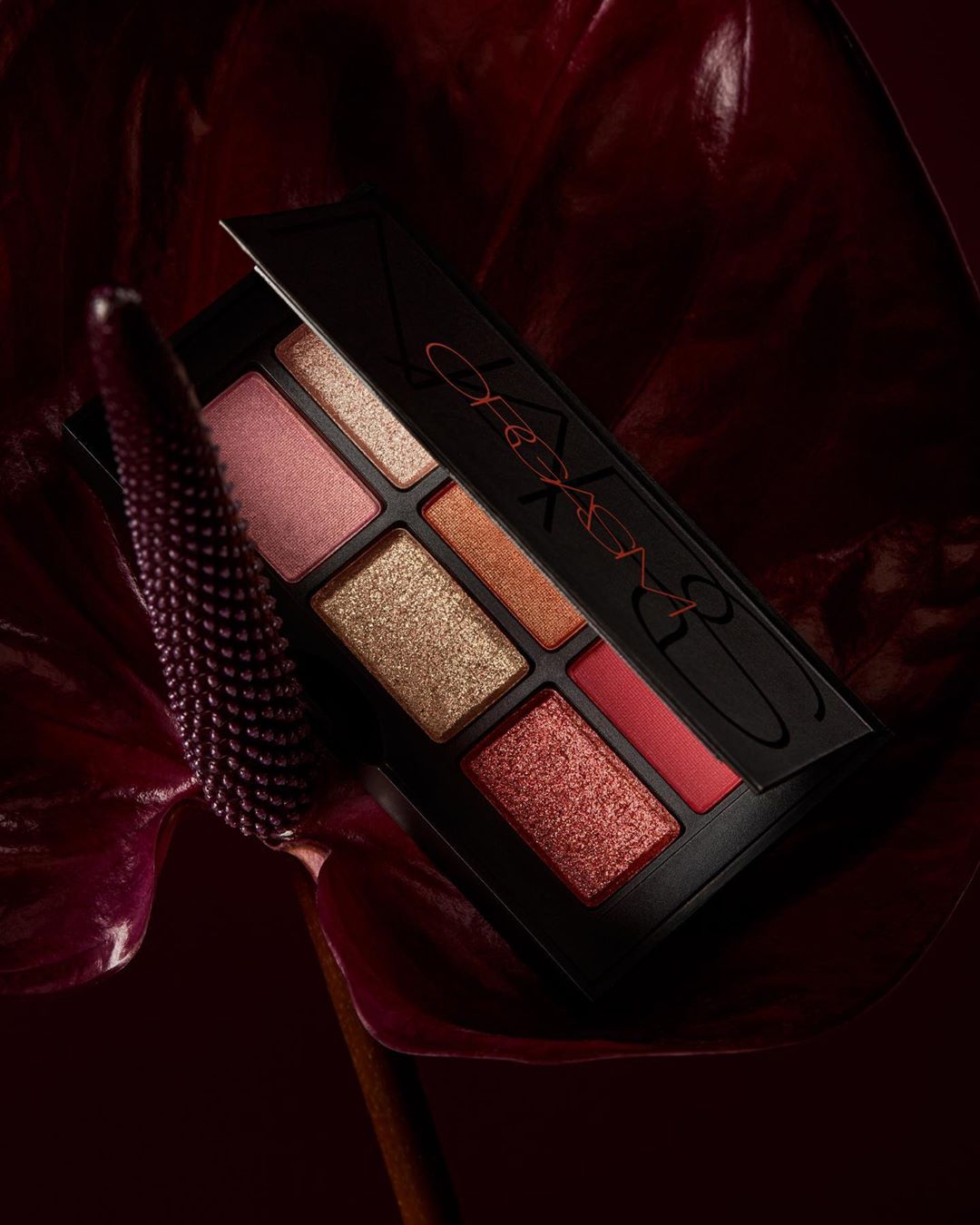Close-up of a NARS eye shadow palette with various shades of pink, gold, and red, displayed with a dark red calla lily.