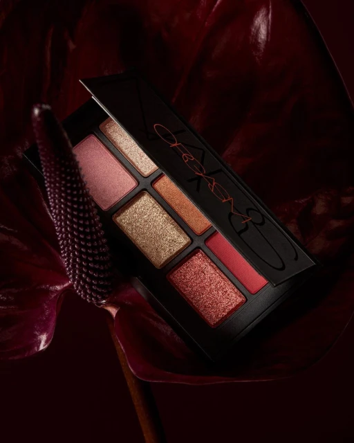 Close-up of a NARS eye shadow palette with various shades of pink, gold, and red, displayed with a dark red calla lily.