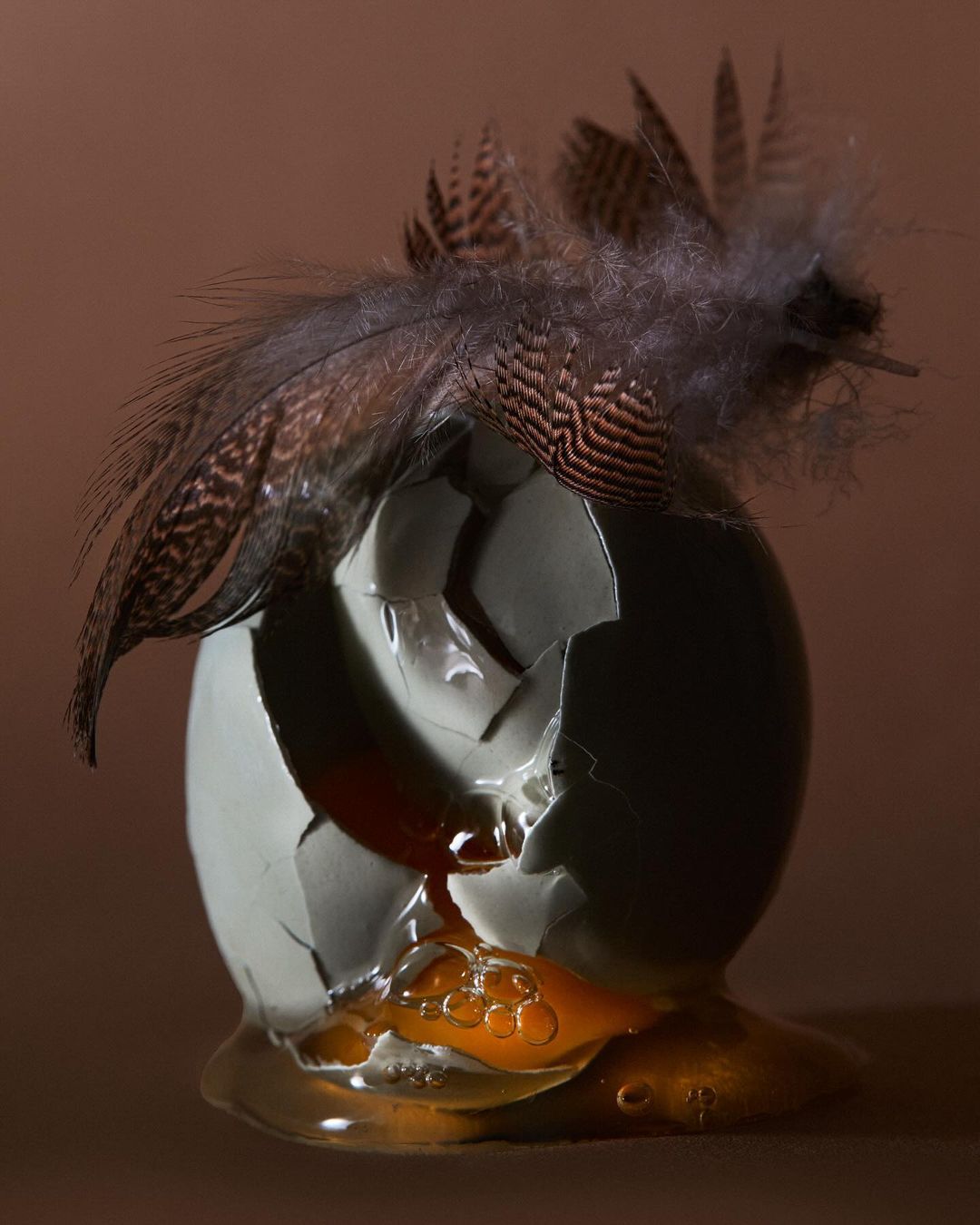 Cracked eggshell with dripping yolk and feathers against a brown background. Food photography with dark and moody lighting.