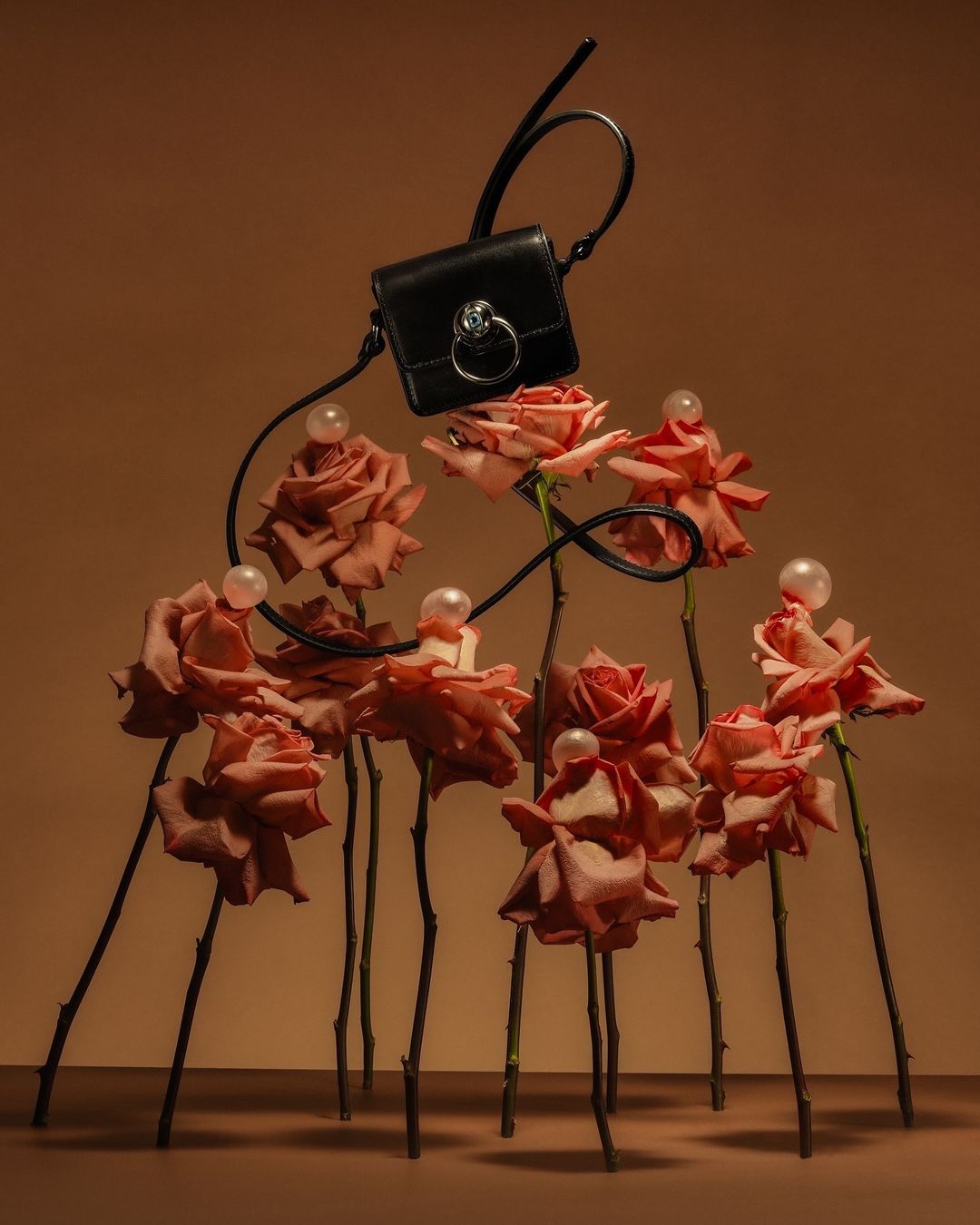 Fashion still life: A black leather purse floats above roses adorned with pearls against a warm brown background, creating an elegant composition.