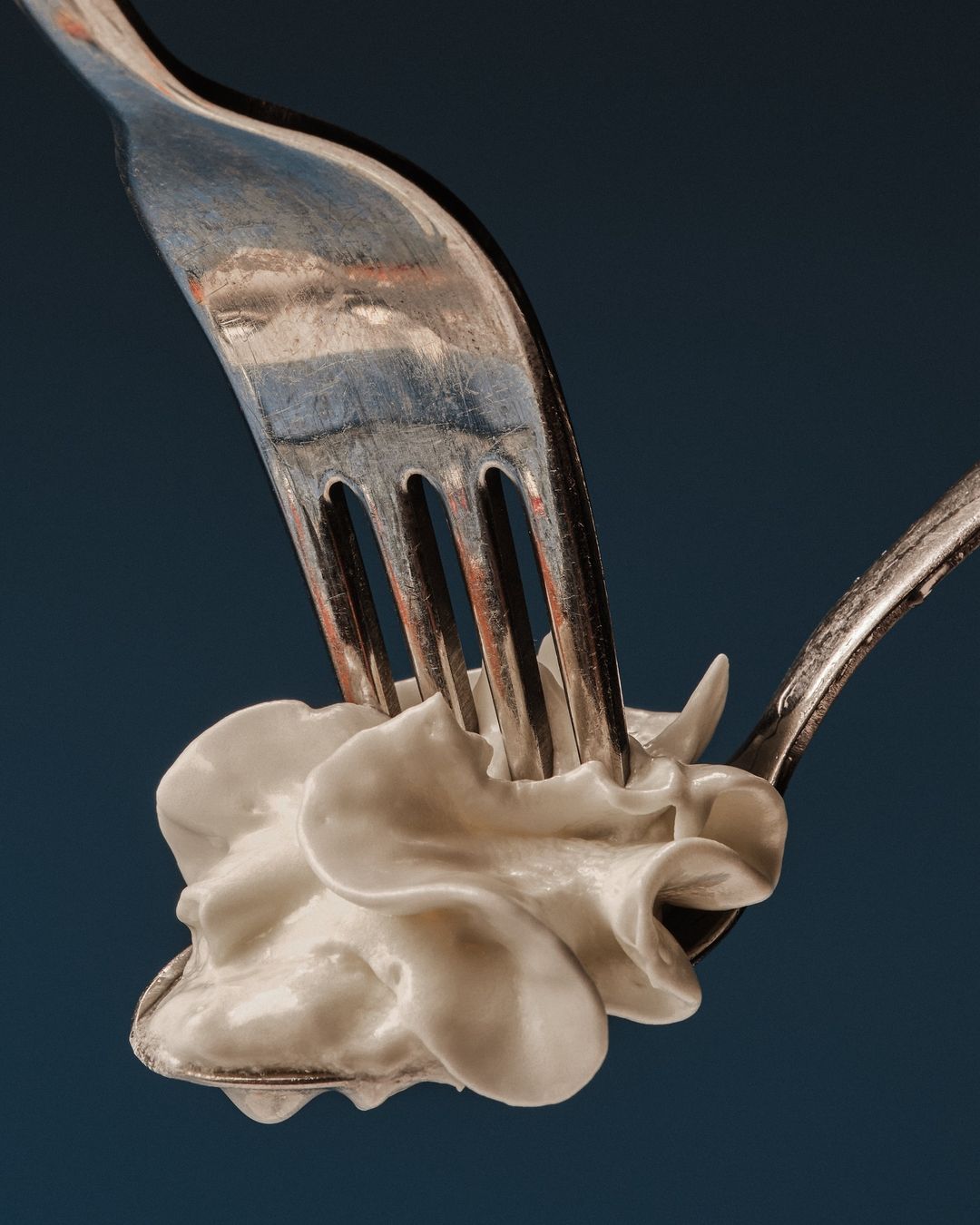 Close-up still life shot of white cream being pierced by a metal fork over a spoon against a dark blue background in studio.