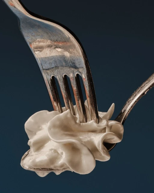 Close-up still life shot of white cream being pierced by a metal fork over a spoon against a dark blue background in studio.