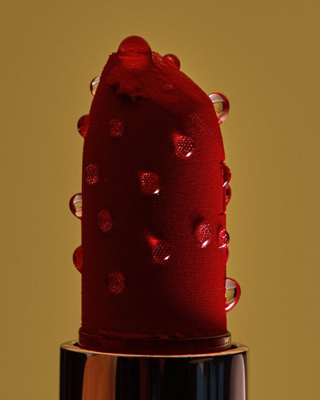Close-up of a red lipstick bullet with water droplets against a light-yellow background in a beauty and cosmetics studio shot.