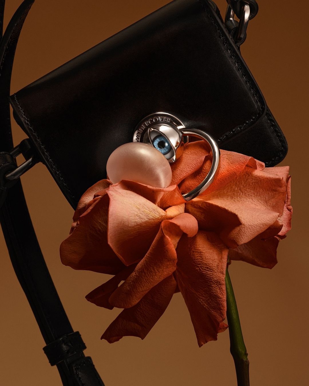 Close-up studio shot of a black purse adorned with a rose, eye detail, and a smooth pink orb against a muted beige background.