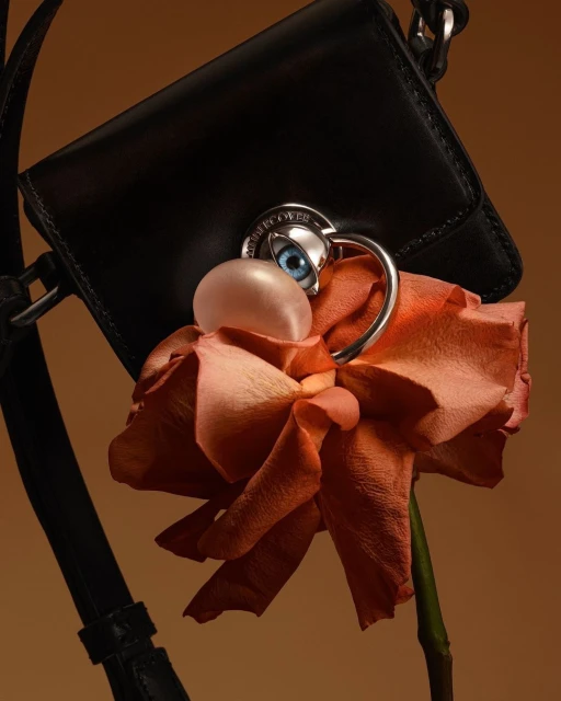 Close-up studio shot of a black purse adorned with a rose, eye detail, and a smooth pink orb against a muted beige background.