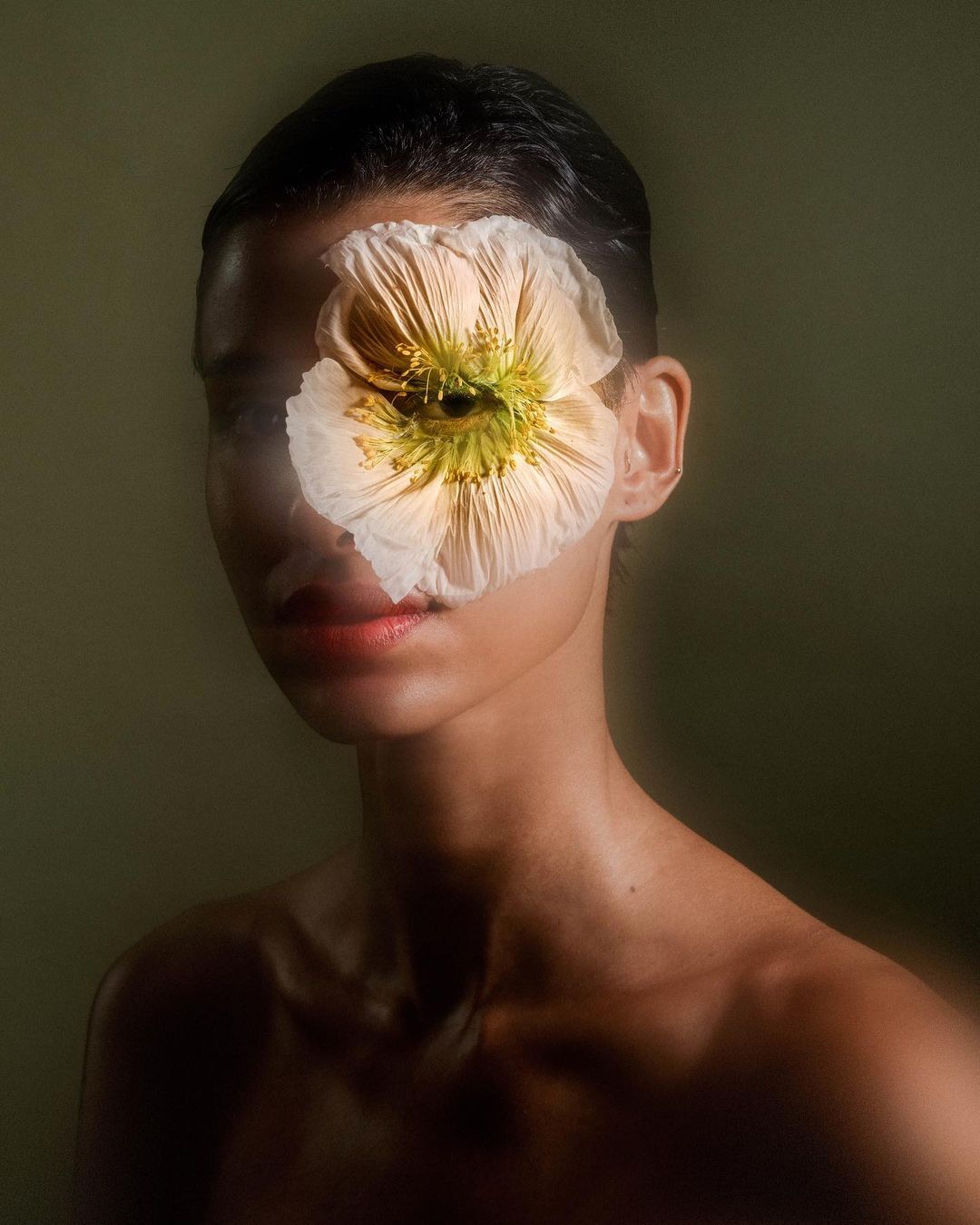 Close-up portrait of a woman with a flower covering her eye, creating a surreal effect, against a muted green background in soft lighting.