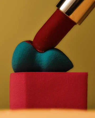 A red lipstick tube presses into a blue makeup sponge atop a pink block against a yellow gradient background, creating a beauty still life.