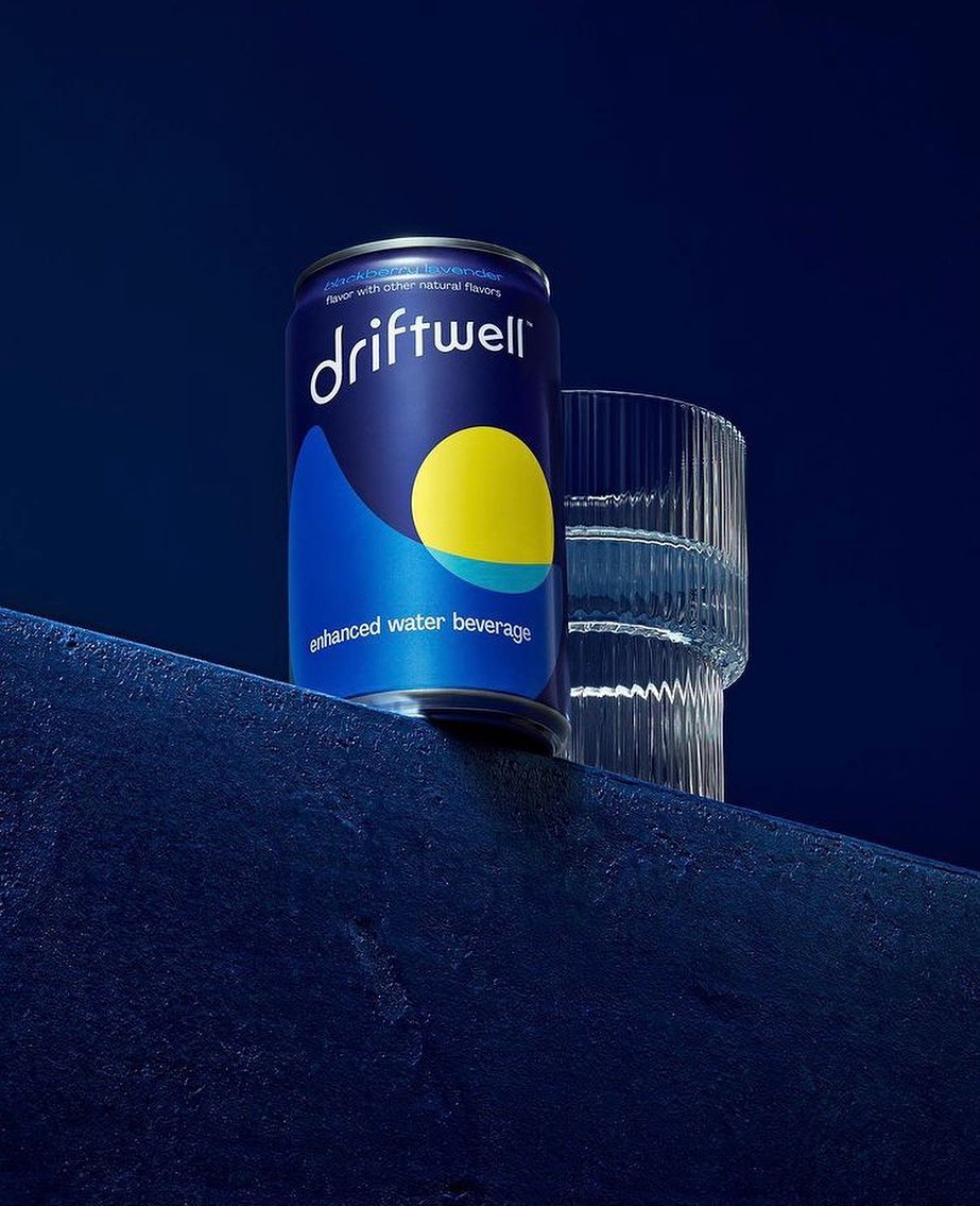 A single can of Driftwell enhanced water beverage is next to a clear glass on a dark blue textured surface against a dark blue background.