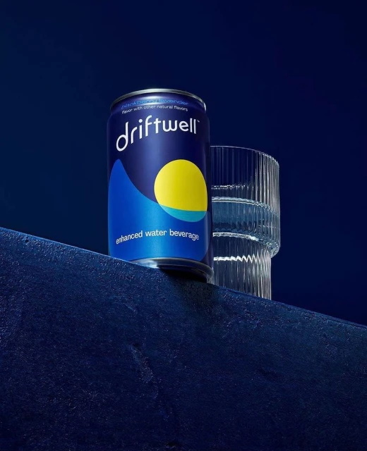 A single can of Driftwell enhanced water beverage is next to a clear glass on a dark blue textured surface against a dark blue background.